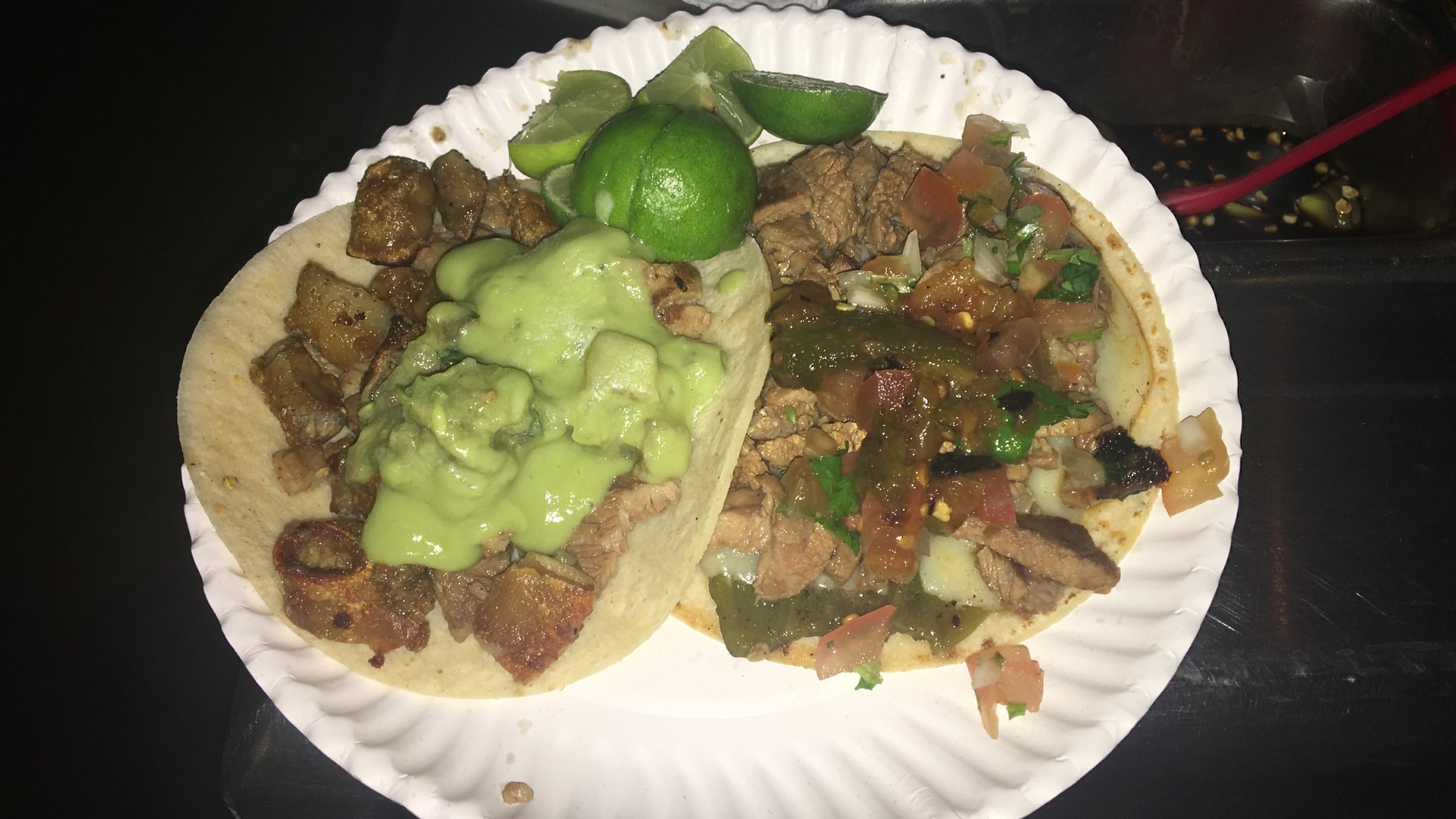 Two beef tacos on a paper plate with salsa, guacamole and lime wedges